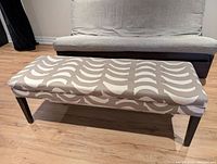 Front view of cushioned bench with fabric upholstery and patterned beige and white design, set against wooden floor and gray sofa.