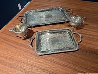 Photo of two silver-toned decorative trays and two matching cups arranged on a wooden surface, showing embossed floral and scroll patterns.