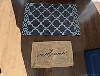 Two doormats laying on wooden floor. Large doormat has geometric pattern, small doormat says 'welcome'.