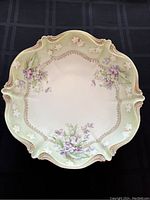 Top view of antique display bowl showing scalloped edges, violet floral motifs, and gilt detailing on a cream and pastel green background.