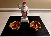 Porcelain Bonhomme figurine standing with raised arm, red hat and sash, base inscribed 'Carnaval Quebec', flanked by two gold gilded flower-shaped Beauceware candle holders on a black surface.