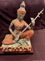 Front view of bronze Thai Gandharva figure holding and playing a string instrument, showing detailed gilded clothing and headdress.