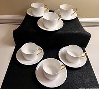 Six bone china cups with gold-plated handles arranged on black cloth, each paired with a saucer.
