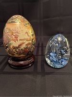 Two decorative eggs shown side by side on a black fabric. Left is the ceramic Satsuma style egg on wooden stand; right is the blue clear glass paperweight egg.