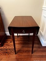 Wood side table with one drawer and drop leaf sides closed. View of front showing drawer with metal ring pull handle and tapered legs.