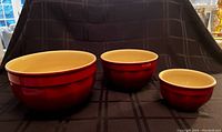 Three ceramic bowls arranged from largest to smallest on a black cloth. Bowls have a red exterior and cream interior.