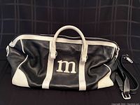Full front view of dark navy and white leather duffle bag showing initial 'm' on the side, resting on black surface.