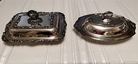 Photo of two vintage silverplated serving dishes side by side on a white tablecloth, showing the rectangular dish on the left with an ornate lid and handle, and the oval dish on the right with a smooth lid and handle.