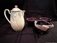 Front angle showing bone china teapot with floral relief design and three ceramic bowls, including two dark purple and one black and white artistic bowl.