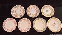 Top-down photo showing all seven vintage china plates, displaying an assortment of floral and decorative patterns including peacock, gold accents, and red floral motifs.