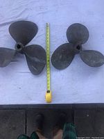 Two vintage bronze or brass boat propellers side by side with a measuring tape showing sizes around 17 and 15 inches