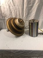 Photo of two ceiling lamps placed side by side on a cloth-covered surface; left is painted metal Deco fixture, right is colored glass cylinder fixture.