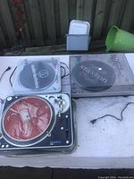 Three DJ turntables on a white surface outdoors, visible power cords, some wear and dust.