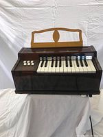 Front view of small brown plastic electric air organ with keyboard and switches, showing keys, function buttons, and music stand.