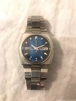 Front view of the watch showing the blue dial, day-date window, and stainless steel case and band.