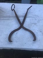 Full view of the large, rusty iron ice pick on a white cloth background showing the crossed hooked ends and loop handles.