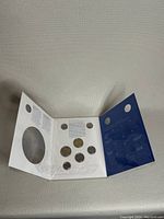 Photo of the tri-fold cardboard holder containing seven coins in mint condition, blue and white background with text.