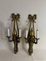 Front view of the pair of antique brass wall lights with two bulbs each, hanging on a wall, showing ribbon detail at top.