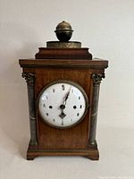 Front view of old German mantel clock showing wooden case with marble-effect columns and white enamel dial with Roman numerals.