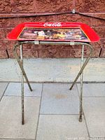 Photo showing full view of Coca Cola table tray with foldable metal stand placed on tiled floor in front of brick wall; tray features vintage Coca Cola artwork and red trim.