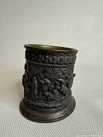 Full view of the antique German brass pot showing cylindrical shape, decorative raised relief figures, and patterned rim and base.
