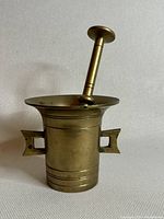 Front view of brass mortar and pestle together showing the shape and features of the mortar, including side handles and decorative lines near base, pestle upright inside mortar.