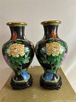 Front view of both mirror image cloisonné vases with colorful floral designs on black background with wooden stands.