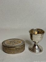 Pair of silver plated pieces including a small cup and an oval trinket box with intricate engraving on the lid and sides.