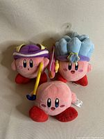 Three Kirby plush toys shown together with different costumes and accessories