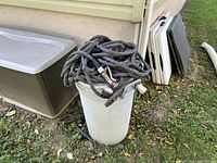 Two black outdoor garden hoses coiled and resting on a white plastic barrel near a house exterior, including visible hose connectors and a spray nozzle.