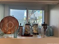 Photo showing assorted ceramic steins with metal lids, large ceramic serving dish, colored shot glasses on tray, decorative glass bottles, wine glass, and clear drinking glass arranged on a white shelf in front of a window.
