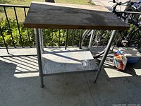 Photo of butcher block table made of wood top and metal legs with lower shelf seen outdoors with surrounding environment showing wear and outdoor use.