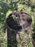 Round metal fire pit with mesh screen cover sitting on grass, showing some rust and leaves inside.