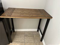 Photo of a wooden high table with black metal legs placed against a wall on a tiled floor.