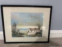 Framed watercolor painting showing a house by a river with natural surroundings and cloudy sky.
