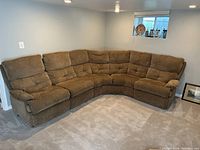 Photo showing the large L-shaped tan sectional sofa in a carpeted room near a small window with several decorative items. All sections feature padded backs and armrests, designed to recline and detach.