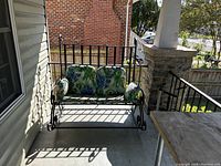 Photo of a black metal porch rocker bench with cushions on a porch beside a stone pillar and railing.