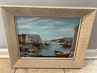 Framed painting showing a coastal scene with boats and dock buildings under a cloudy sky.