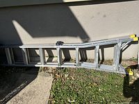 Side view of an extendable aluminum ladder with adjustment mechanism and yellow end caps lying on the ground outside against a wall.