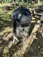 Outdoor vertical drum smoker with a domed lid, front access door, and vent control on top. Shown standing on grass outdoors with sunlight and nearby outdoor furniture.