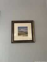 Framed artwork showing sandy mountain landscape with clouds, mounted with a cream matte inside a decorative dark wooden frame with gold details.