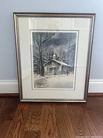 Framed print of a stone building in the snowy woods titled 'Country School' by Richard Bollinger, set against a pale wall and hardwood floor.