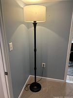 Tall black metal floor lamp with beige fabric drum shade lit in a corner of a room with light blue walls and beige carpet.