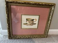 Photo of the framed tea cup print showing the ornate golden frame and pink matting with central print.