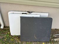 Three folding tables leaning against wall outdoors showing white and dark tops with wear and stains.