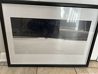 Framed black and white photograph showing a seaside scene at night with people walking along the shore. The frame is black and the image is matted with white border.