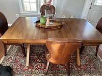 Dining table and chairs shown from above and side angle, displaying table surface, legs, and leather chairs in a well-lit room with a patterned rug underneath.