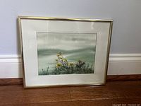 Framed artwork showing yellow flowers against a grey sky background and the thin gold frame on a wooden floor surface.