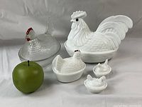 Front view of the 5 glass chicken casserole dishes displayed with a green apple for size reference. Shows large milk white rooster, 3 milk white hens, and 1 clear glass hen with red comb.