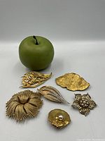 Photo with green apple for size reference showing all six gold-tone brooches with various floral and abstract designs including pearl and rhinestone accents.